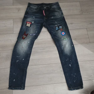Mörkblå jeans med patches och stänk - Unika mörkblå jeans med coola patches och färgstänk framtill. Jeansen har klassisk femficksmodell, smal passform och slitna detaljer. Flera broderade märken och tryck ger en streetig vibe. Perfekta för dig som vill sticka ut med din stil.