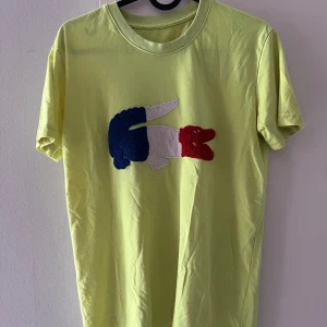 Gul Lacoste t-shirt med krokodilmärke - Gul t-shirt från Lacoste med stort broderat krokodilmärke i blått, vitt och rött på bröstet. Klassisk rund hals och korta ärmar. Perfekt för dig som vill sticka ut med en färgglad och ikonisk look.