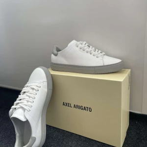 Vita sneakers från Axel Arigato - Stilrena vita sneakers från Axel Arigato med grå detaljer och platt sula. Skorna har minimalistisk design, snörning och är tillverkade i skinn. Perfekta för dig som gillar en clean och modern look. Pris kan diskuteras samt andra frågor. 