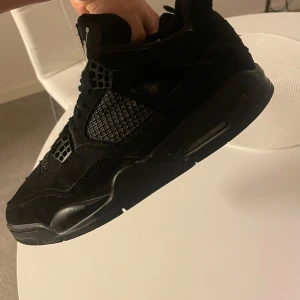 Svarta Nike Air Jordan 4 sneakers - Säljer ett par helsvarta Nike Air Jordan 4 sneakers med snygga meshdetaljer på sidorna och klassisk Jumpman-logga på hälen. Skorna har snörning, platt sula och är tillverkade i mocka och syntet. Perfekta för dig som gillar en clean och sportig look.