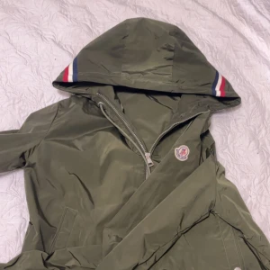 Grön vindjacka från Moncler med huva - Snygg grön vindjacka från Moncler med huva och dragkedja. Jackan har Moncler-logga på ärmen och coola röd, vit och blå detaljer på huvan. Perfekt för blåsiga dagar och enkel att matcha med streetwear. Fickor med tryckknappar och justerbar huva.