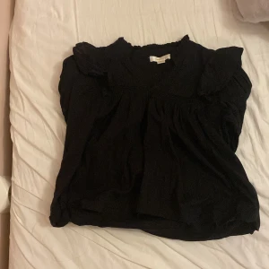 Svart blus med volang från Gina Tricot - Supersöt svart blus från Gina Tricot med volangdetaljer vid axlarna och smockade ärmslut. Blusen har v-ringning och rynkade partier som ger en luftig och flowy känsla. Perfekt att styla med jeans eller kjol för en trendig look. Storlek 146/152