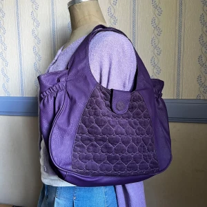 Lila axelväska med hjärtdetalj - En stor lila axelväska från PiGi med ett  guldfärgat hjärtformat dragkedjesmycke och flera fack. En extra liten väska tillkommer, är trasig i fästet på kedjan men går att byta ut 🩷