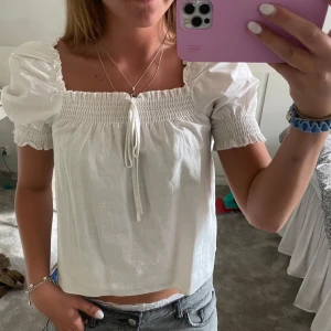 Vit blus med puffärm och smock - Supersöt vit blus med puffiga korta ärmar och smock upptill. Blusen har fyrkantig ringning och en liten knytning framtill. Den är lätt och luftig, perfekt för varma dagar. Passar dig som gillar romantisk och trendig stil.