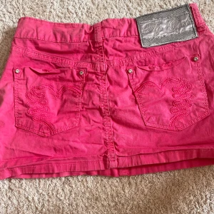 Rosa jeanskjol - Snygg rosa jeanskjol från Fly Girl med broderade detaljer på bakfickorna och silvrig logga framtill. Klassisk femficksmodell med knapp och dragkedja. Perfekt för dig som vill sticka ut med färg och coola detaljer. 2000 tal stil och unikt! 