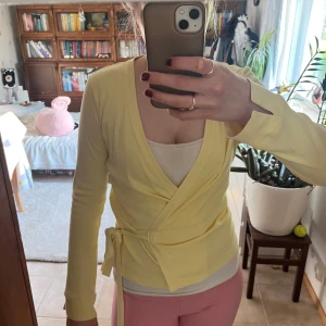 Gul omlottkofta med knytband - Söt ljusgul omlottkofta med långa ärmar och knytband i midjan. Koftan har en djup v-ringning och är i ett mjukt, stretchigt material som känns skönt mot huden. Perfekt att slänga över ett linne för en fräsch och vårig look.