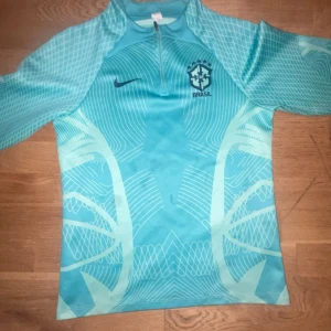 Turkos Brasil Nike fotbollströja - Snygg turkos Brasilien fotbollströja från Nike med halv dragkedja och coola grafiska mönster i ljusblått. Perfekt för dig som vill sticka ut på planen eller på läktaren. Priset kan diskuteras 