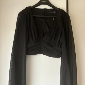Svart croppad blus PrettyLittleThing - Säljer en svart croppad blus från PrettyLittleThing med transparenta, långa ärmar i mesh. Toppen har djup v-ringning, rynkade detaljer framtill och en figurnära passform. Perfekt för dig som vill ha en trendig och stilren look.