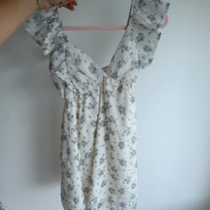 Blommig kortklänning med volang - Supersöt kortklänning i ljus färg med blått blommönster. Klänningen har breda volangprydda axelband, v-ringning och en rosett i ryggen. Perfekt för sommarens alla tillfällen