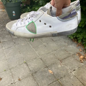 Philippe Model vita sneakers med camo-detalj - Snygga vita sneakers från Philippe Model med grå mockadetaljer och cool kamouflagemönstrad häl i grönt, blått och beige. Skorna har snörning, platt sula och en unik grön kant runt loggan på sidan. Perfekta för dig som gillar streetstyle med en twist.