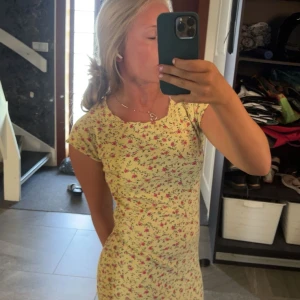 Gul blommig kortklänning - Supersöt gul kortklänning med små blommor i rosa och grönt. Klänningen har korta ärmar och en figurnära passform som ger en fräsch sommarkänsla. Perfekt för dig som gillar en feminin och lekfull stil.