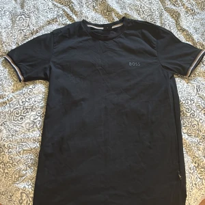 Svart t-shirt från BOSS med logga - Svart t-shirt från BOSS med diskret logga på bröstet och liten etikett i nederkant. Klassisk rund hals och korta ärmar med snygga randiga detaljer vid ärmsluten. Skön och mjuk bomullskvalitet, perfekt för en clean och stilren look.