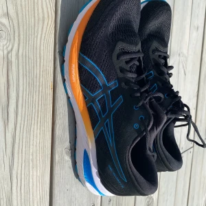 Svarta Asics sneakers med blå detaljer - Snygga svarta Asics sneakers med blå logga och detaljer, samt orange och vit sula. Ovandelen är i mesh för bra andningsförmåga och skon har snörning. Perfekta för dig som gillar att springa eller träna och vill ha en sportig look.