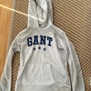 Vit  hoodie från GANT med tryck - Snygg vit  hoodie från GANT med blått broderat logotyp-tryck och stjärnor på bröstet. Klassisk känguruficka framtill och huva. Perfekt för chill dagar eller när du vill ha en sportig look.