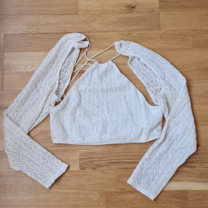 Offwhite topp i spets Bershka - Superfin off white croppad topp från Bershka med långa ärmar och spetsdetaljer. Toppen har knytning i nacken och öppen rygg, samt virkade detaljer som ger en boho-vibe. Perfekt för sommaren och festivaler!