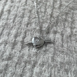 Flèche d’Argent - Silver Hjärthalsband med Pil - Silverpläterat halsband i stainless steel med hjärta och pil, dekorerat med glittrande stenar. Justerbar längd 44 + 4 cm. En tidlös symbol för kärlek med modern elegans