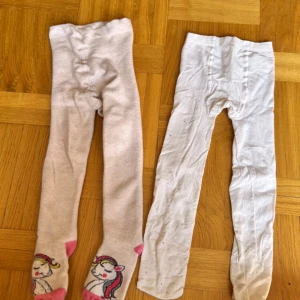 2-pack vita och ljusrosa leggings 98/104 - Två par leggings i barnstorlek 98/104. Ena paret är ljusrosa med söta enhörningar och rosa detaljer vid fötterna, det andra paret är helvita. Båda är mjuka och stretchiga, perfekta för lek och vardag.