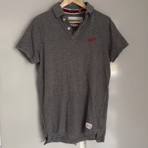 Grå pikétröja från Superdry Medium - Klassisk grå pikétröja från Superdry med korta ärmar och krage. Tröjan har knappar vid halsen och röd broderad logga på bröstet. Tillverkad i mjuk bomull, perfekt för en avslappnad och stilren look.
