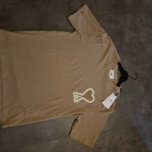 Beige t-shirt från Ami med hjärtlogo - Snygg beige t-shirt från Ami med stor broderad hjärtlogo på bröstet och diskret Ami-broderi i nacken. Klassisk passform, rund hals och korta ärmar. Tillverkad i mjuk bomull för en clean och stilren vibe. 