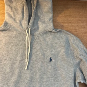 Grå hoodie från Polo Ralph Lauren - Klassisk grå hoodie från Polo Ralph Lauren med blå broderad logga på bröstet. Tröjan har huva med snörning, känguruficka och långa ärmar. Perfekt för en chill och stilren look. Tillverkad i mjuk bomullsmix för extra komfort.