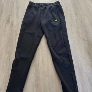 Svarta mjukisbyxor från Lyle & Scott - Marinblåa mjukisbyxor från Lyle & Scott med klassisk gul logga på ena benet. Byxorna har dragsko i midjan och smal passform. Perfekta för chill eller träning, tillverkade i mjukt material som känns skönt mot huden. 