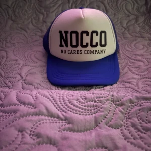 Blå och vit NOCCO keps - Snygg keps från NOCCO med vit frontpanel och blå skärm samt baksida. Stor svart text framtill med loggan 'NOCCO No Carbs Company'. Perfekt för dig som gillar sportig och trendig stil.