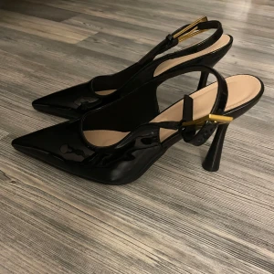 Svarta lackpumps med spetsig tå - Snygga svarta pumps i lack med spetsig tå och hög klack. Skorna har öppen häl med justerbar rem och guldfärgat spänne som detalj. 