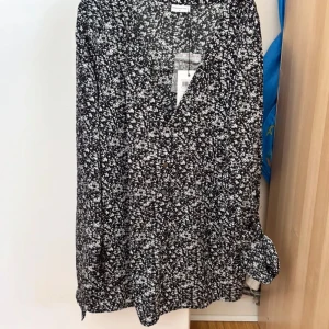 Svartvit blommig klänning JDY - Svartvit kortklänning från Jacqueline de Yong med småblommigt mönster, v-ringning och knappar framtill. Långa ärmar med lätt puff och smalare ärmslut. Luftig passform, perfekt för en trendig look.