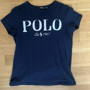 Mörkblå t-shirt från Polo Ralph Lauren - Mörkblå t-shirt från Polo Ralph Lauren med vit broderad POLO-logga och texten 'Est. 1967' framtill. Klassisk rund hals och korta ärmar. Skön passform i mjuk bomull, perfekt till jeans eller shorts. Storlek m men skulle säga att den passar S
