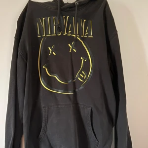 Svart Nirvana hoodie med tryck - Svart hoodie med Nirvana-tryck i gult och stort smileyface på bröstet. Klassisk känguruficka framtill, huva med snörning och långa ärmar. Perfekt för dig som gillar bandmerch och vill ha en avslappnad stil. Finns ett litet märke som syns på bild annats helt felfri 