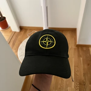 Mörk grön keps från Stone Island - Mörk grön keps från Stone Island med klassisk böjd skärm och justerbar rem bak. Framtill sitter det ikoniska Stone Island-emblemet i gult och svart broderat. Kepsen är tillverkad i bomull och har en clean, sportig look som passar perfekt till streetwear.