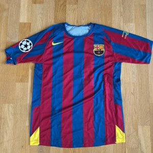 FC Barcelona Ronaldinho 10 Nike tröja S 05-06 - Klassisk FC Barcelona matchtröja med Ronaldinho 10 på ryggen. Röd och blå vertikala ränder, officiellt klubbmärke på bröstet och Champions League-märke på ärmen. Tillverkad i lätt och ventilerande polyester, perfekt för fotboll eller samling. (Aldrig använd)