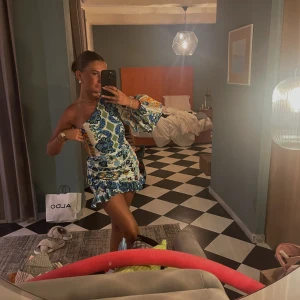 Blommig one shoulder kortklänning - Superfin kortklänning med färgglatt blommönster i blått, grönt och orange. One shoulder-modell med lång ärm på ena sidan och volangdetalj nertill. Sitter tajt över kroppen. Linne blanding i storlek XS