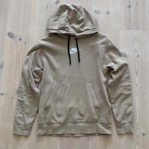 Beige Nike hoodie  - Snygg beige hoodie från Nike med svart logga på bröstet och vita band med små Nike-loggor längs ärmarna. Den här används ett par gånger men det märks knappt så den är nästan som ny