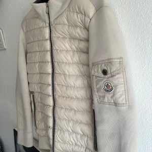 Beige pufferjacka från Moncler - Snygg beige pufferjacka från Moncler med quiltad framsida och ärmficka med logga. Jackan har dragkedja framtill, ribbade muddar och två fickor med dragkedja. Perfekt för dig som vill ha en clean och stilren look med exklusiv känsla.