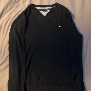 Svart v-ringad tröja Tommy Hilfiger - Svart v-ringad tröja från Tommy Hilfiger med diskret logga på bröstet. Tröjan har långa ärmar och ribbade muddar vid ärmslut och nederkant. Perfekt för dig som gillar stilrena och klassiska plagg.