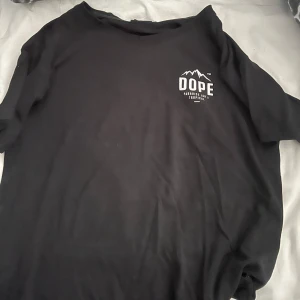 Svart t-shirt från Dope med tryck - Svart t-shirt från Dope med stort vitt tryck på ryggen och mindre logga på bröstet. Klassisk rund hals och korta ärmar. Materialet är mjuk bomull och passformen är normal. Perfekt för dig som gillar streetwear och tydliga prints.