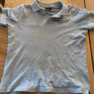 Ljusgrå pikétröja från Peak Performance - Säljer en ljusgrå pikétröja från Peak Performance med klassisk krage och knappar framtill. Tröjan har korta ärmar och diskret logga broderad på bröstet. Perfekt för en avslappnad och sportig stil.