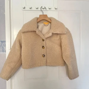Beige teddyjacka med knappar - Säljer en beige teddyjacka med stora slag och knappar framtill. Jackan är croppad och har lång ärm. Perfekt för dig som vill ha en mysig och trendig look. Ull jacka perfekt för våren och svalare väder. 