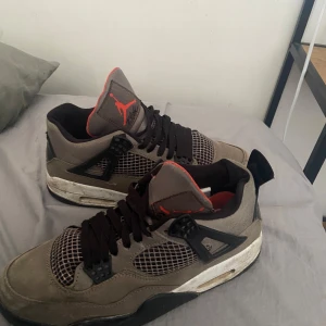 Nike Air Jordan 4 sneakers i grått och svart - Säljer ett par Nike Air Jordan 4 sneakers i grå mocka med svarta och röda detaljer. Skorna har klassisk mesh på sidorna, svart snörning och vit sula med synlig Air-enhet. Röd insida och Jumpman-logga på plösen och hälen.