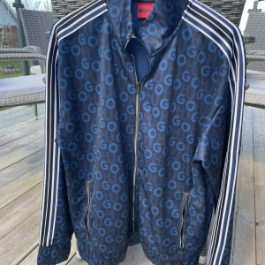 Blå mönstrad track jacket från HUGO - Säljer en blå track jacket från HUGO med all-over logomönster i blå nyanser. Jackan har vita ränder längs ärmarna, dragkedja framtill och två fickor med dragkedja. Perfekt för en sportig och trendig look.