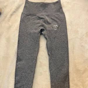 Grå scrunch leggings från Peach - Gråa leggings med hög midja från Peach. Byxorna har scrunch-detalj bak och är tillverkade i stretchigt material som passar perfekt för träning. Liten broderad logga framtill och texten 'peach' bak på linningen.