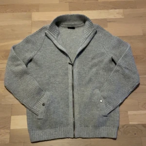 Grå stickad kofta  - Säljer en grå stickad kofta med dragkedja och hög krage. Koftan har långa ärmar, ribbade muddar och fickor med knappdetalj. Inga defekter. Storlek L men skulle kunna passa M också