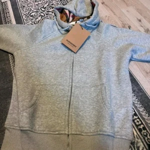Grå hoodie från Burberry med dragkedja - Säljer en grå hoodie från Burberry med dragkedja och klassiskt rutigt foder i huvan. Tröjan har fickor framtill och ribbade muddar. Perfekt för en avslappnad och stilren look. Helt ny och oanvänd. Storlek 38/M men liten i storlek.