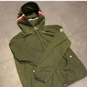 Grön vindjacka med huva från Moncler - Snygg grön vindjacka från Moncler med huva och dragkedja. Jackan har en vit och röd rand på huvan samt Moncler-logga på ärmen. Perfekt för blåsiga dagar och enkel att matcha med olika outfits.