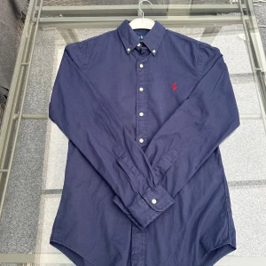 Ralph Lauren skjorta slim fit S marinblå - (ÖPPPEN FÖR BUD)                                           Säljer en klassisk skjorta från Ralph Lauren i storlek S, slim fit-modell. Marinblå färg, mjukt och tåligt oxford-tyg, button-down-krage och röd broderad Polo-logga på bröstet. Perfekt till både vardag och fest – passar till jeans, chinos eller under en kavaj.  Skjortan är i mycket fint skick, utan fläckar eller skador. Kommer från ett djur- och rökfritt hem. Snabb leverans!  