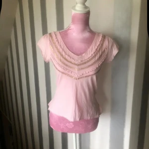Ljusrosa blus med volang och guldiga detaljer - Söt ljusrosa blus med v-ringning och korta ärmar. Framsidan har dekorativa guldiga band och volangdetaljer, samt en volangkant nedtill. Perfekt för dig som gillar romantisk och feminin stil.