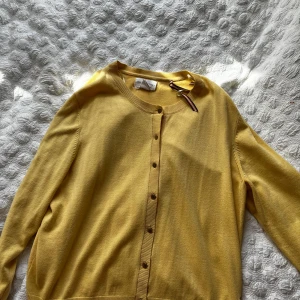 Gul kofta från Holly & Whyte - Säljer en söt gul kofta från Holly & Whyte. Koftan har rund halsringning, knappar framtill och långa ärmar. Perfekt att slänga över en topp för en färgklick i garderoben!