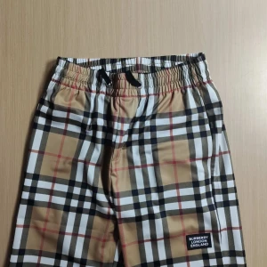 Burburry shorts - Snygga byxor med klassiskt rutigt mönster i beige, svart, vitt och rött. Byxorna har resår i midjan med snörning och en liten Burberry-logga på benet. Perfekta för en trendig och avslappnad stil.