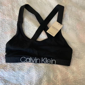 Svart sport-bh från Calvin Klein - Svart sport-bh från Calvin Klein med bred resår och logga framtill. Axelbanden är korsade i ryggen för extra stöd. Perfekt för träning eller som bekväm topp till vardags.